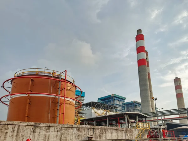 Büyüleyici düşük açılı çekim bulutlu bir gökyüzünün altında endüstriyel bir kompleks sergiliyor. Etkinlikte önde gelen depolama tankları ve yüksek bacalar yer alıyor, enerji ve üretim süreçlerini vurguluyor