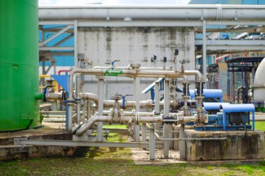 Birçok kapakçığa ve büyük bir yeşil depolama tankına sahip endüstriyel borular bir açık hava işleme fabrikasının ön planına hakim. Mavi motorlar yoğun bir çalışma ortamını gösteren beton kaidelerde görülebilir.
