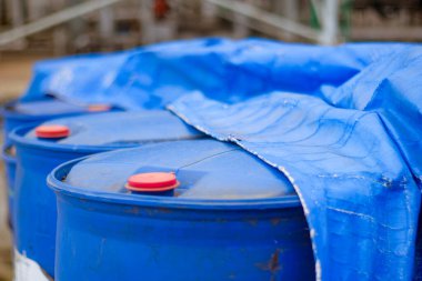 Parlak kırmızı kapaklı çoklu mavi plastik depolama fıçıları açık hava endüstriyel ortamında mavi koruyucu muşamba ile kısmen kaplıdır.