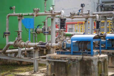 Büyük yeşil bir tank yakınında, endüstriyel bir fabrikada çalışan paslanmaz çelik borulardan oluşan karmaşık bir ağ kontrol vanaları ve elektrik pompaları.