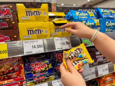 Parlak ışıklı bir market şekerleme bölümünden bir paket çikolatalı şekere uzanan müşterinin elini kapatıyoruz.