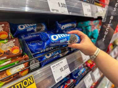 Kişi, marketin koridorundan çeşitli bisküvi ürünleri ve fiyat etiketleriyle dolu mavi bir sandviç paketi seçer.