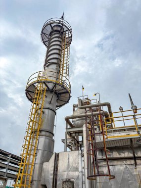 Endüstriyel fabrika, bulutlu bir gökyüzü altında yoğun bir üretim ortamının altını çizen belirgin bir baca ve karmaşık bir boru hattına sahiptir.