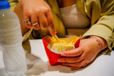 Kırmızı bir karton patates kızartmasına uzanan ve yanında bir şişe su olan ellerin yakın plan çekimi. Günlük yemek konsepti.