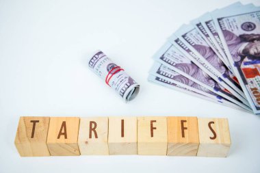 Wooden cubes spelling 'Tariffs' next to a fan of US dollar banknotes, representing economic policy, trade, and finance.