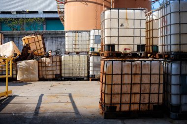 Endüstriyel depolama alanında, çeşitli aşınma durumlarında çok sayıda orta büyüklükteki konteynır büyük fabrika tankları ve parlak güneş ışığı altındaki binalara karşı dizilmiş durumda.