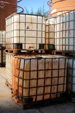 Dışarıdaki paletlerin üzerinde paslı metal çerçeveler ile kaplanmış beyaz plastik tankları olan çoklu yıpranmış sıvı depolama konteynerlerini gösteren endüstri sahnesi.