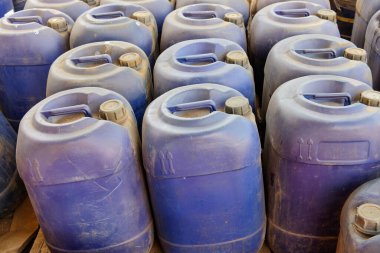 Mavi plastik bidonlar, endüstriyel bir depolama alanında sıkıca bir araya toplanır ve yıpranmış yüzeylerinde görünür toprak ve çizikler bulunur.