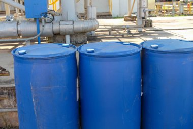 Beyaz kapaklı mavi plastik konteynırlar, arka planda depolama amaçlı güpegündüz borular ve makinelerle açık bir sanayi bölgesinde düzenlenir.