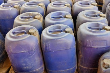 Mavi plastik bidonlar, endüstriyel bir depolama alanında sıkıca bir araya toplanır ve yıpranmış yüzeylerinde görünür toprak ve çizikler bulunur.