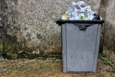 Duvarın köşesinde, gri plastik geri dönüşüm kaplarından toplanıp boşaltılmayı bekleyen dolu çöp kutuları var..