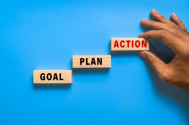 Erkek eli hedefi, planı, eylem sözcüklerini mavi arkaplanda izole edilmiş tahta bloklara koyar