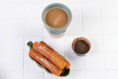 Çikolata soslu çıtır churro ve bir fincan kahve. Temiz beyaz bir masaya dizilmiş..