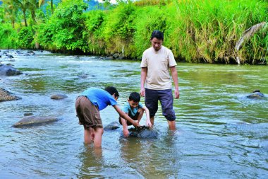 Bir baba, çocuklarına nehir kıyısındaki kayaları ve sığ suları keşfederken yardım eder..