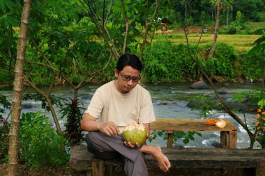 Kırsal bir ortamda genç hindistan cevizi suyunu tutan ve içen bir adam doğal olarak ferahlatıcı bir anın tadını çıkarıyor..