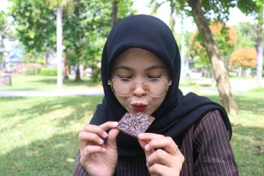 Siyah tesettürlü ve gözlüklü Endonezyalı genç bir kadının parkta çikolatalı gofreti ısırmaya hazırlanırken komik bir surat ifadesi..