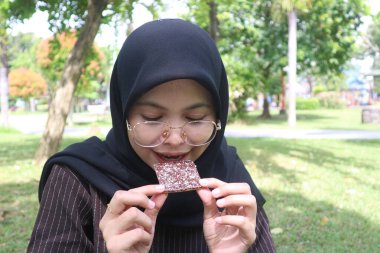 Siyah tesettürlü ve gözlüklü mutlu Endonezyalı bir kadının yakın çekimi. Elinde tuttuğu çikolatalı gofrete gülümsüyor..