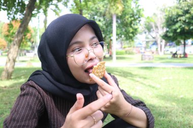 Tesettürlü genç bir kadın çikolata dolu bir bisküviyi ısırır, dışarıda parkta dinlenirken lezzetli bir atıştırmanın tadını çıkarır..