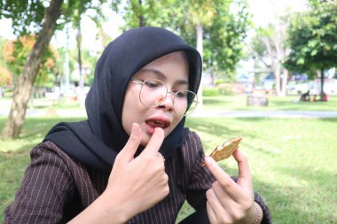 Tesettürlü ve gözlüklü bir kadın çikolata dolu bir bisküviye bakıyor, dışarıda parkta dinlenirken tatlı bir anın tadını çıkarıyor..