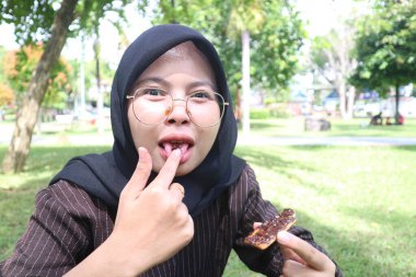 Şakacı, tesettürlü genç bir kadın parmağından çikolata yalıyor. Parkta bisküvi yerken keyifli bir ifade..