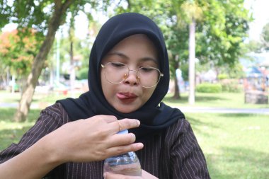 Başörtüsü ve gözlüklü genç bir kadın, neşeli bir ifadeyle, açık havada bir su şişesi tutuyor, atıştırmalıktan sonra susuz kalmaya hazır..