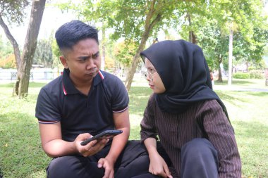 Genç bir Asyalı çift, başörtüsü takan bir erkek ve bir kadın, dışarıda akıllı telefonlarıyla oynarken şüpheli bir şekilde birbirlerine bakıyorlar. Fotoğraf güneşli bir günde parkta çekildi..