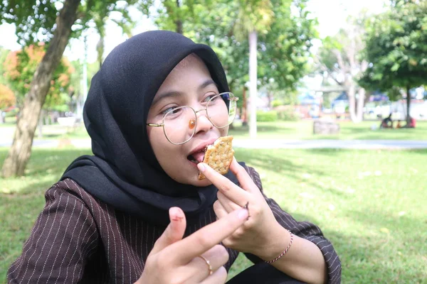 Tesettürlü genç bir kadın çikolata dolu bir bisküviyi ısırır, dışarıda parkta dinlenirken lezzetli bir atıştırmanın tadını çıkarır..