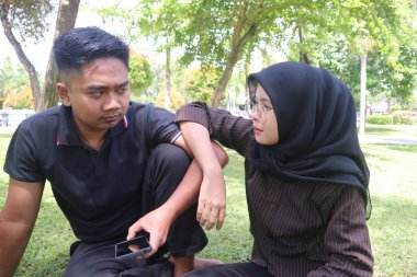 Endonezyalı bir çift parkta çimlerin üzerinde oturuyor, konuşuyor ve birbirlerine bakıyor. Adam siyah polo gömlek giyiyor ve kadının siyah tesettüre ve gözlüğe sahip..