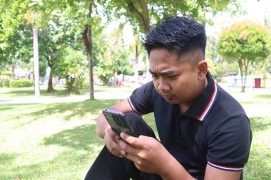 Endonezyalı genç bir adam parkta çimlerin üzerinde tek başına oturuyor, akıllı telefonunu yüzünde hafif bir somurtmayla tutuyor..