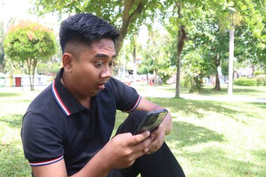 Şok olmuş genç Endonezyalı bir adam parkta çimlerin üzerinde oturuyor, akıllı telefonunu tutuyor ve ona geniş gözlerle bakıyor. Çizgili çizgili siyah bir polo tişört giyiyor..