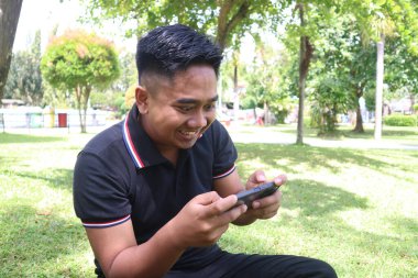 Genç adam gülümsüyor ve mutlu bir şekilde telefonunu parkta kullanıyor..