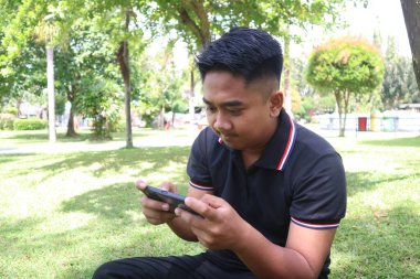 Yeşil bir parkta akıllı telefonuyla oyun oynamaya dalmış genç bir adam. Bağlantı, teknoloji ve eğlence..