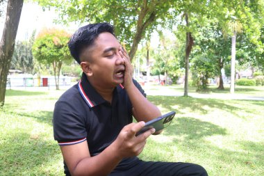 Genç bir adam parktaki telefonuna bakarken yüzünü kapatarak öfkeyle ya da şaşkınlıkla tepki verir..