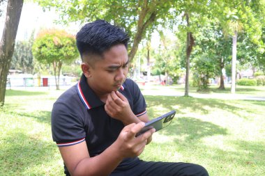 Kafası karışmış genç bir adam, parkta telefonuna bakıyor..