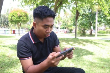 Parkta telefonuna bakan şaşkın ve şaşkın bakışlı genç bir adam..