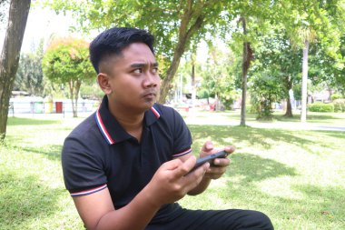 Genç adam bir parkta telefonunu tutarken dikkatle başka tarafa bakıyor..