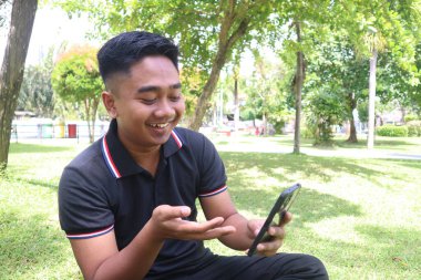 Genç bir adam parkta bir video görüşmesi sırasında elini telefona doğrulturken gülümsüyor..