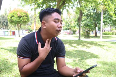 Acılı bir suratı olan genç bir adam. Parkta telefonunu tutarken boğazını tutuyor..