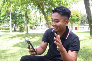 Parkta gülümseyen ve telefonuna el sallayan genç adam. Video çağrısı, teknoloji ve dostça selamlaşma..