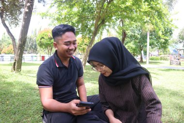 Güneşli bir parkta çimlerin üzerinde oturan mutlu genç bir çift birlikte cep telefonuna bakıyor, açık havada buluşmanın tadını çıkarıyor ve anı paylaşıyor..