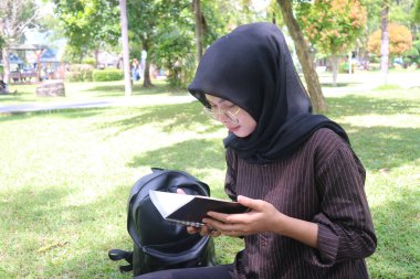 Tesettürlü genç bir kadın parktaki çimlerin üzerinde sırt çantasının yanında defter okuyor. Açık hava çalışmasını, odaklanmayı ve doğa eğitimini simgeliyor..