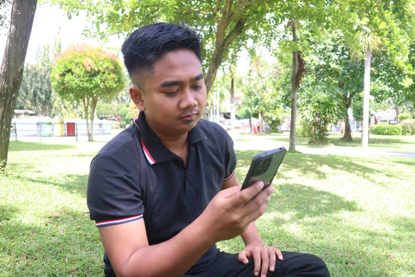 Siyah polo tişörtlü genç adam parkta telefonuna bakıyor..