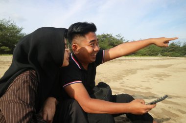 Kumsalda mutlu bir çift oturuyor, gülümsüyor ve adam uzaktaki bir şeye heyecanlı bir şekilde işaret ediyor, güneşli ve neşeli bir sahil anının tadını çıkarıyor..