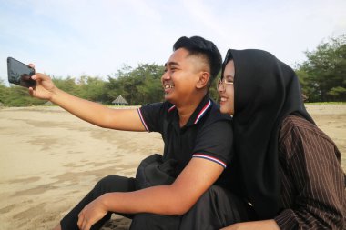 Güneşli bir sahilde selfie çeken gülümseyen bir çift, keyifli bir anın tadını çıkarıyor ve deniz kenarında anılarını yakalıyor..