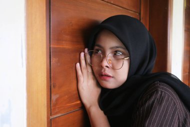 Tahta bir kapıdan gizlice dinleyen tesettürlü, tesettürlü ve gözlüklü, meraklı bir kadın merak ve gizlilik anı yakalıyor..