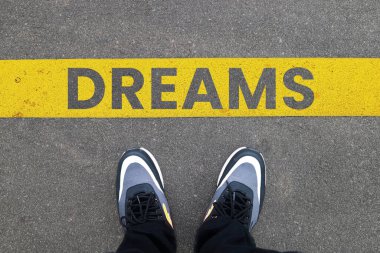 DREAMS metniyle sarı çizginin arkasında asfalt üzerinde duran spor ayakkabılarının üst görüntüsü. Gelecekteki hedefler kavramı.