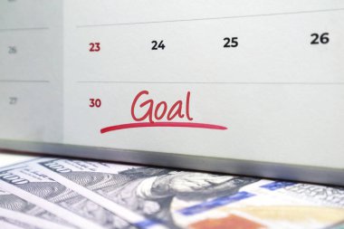 Beyaz spiral takvimde kırmızı mürekkeple yazılmış Goal takvimi dağınık arkaplanda 100 dolarlık banknotlar halinde duruyor..