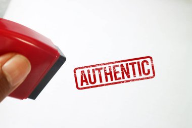 Temiz beyaz bir arka planda kırmızı bir AUTHENTIC mürekkep izinin yanında kırmızı kauçuk bir damga tutuyor. Orijinal ürünler ve resmi doğrulama için kavramsal resim.