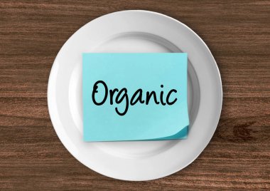 Ahşap bir arka planda beyaz bir tabakta organik yazı olan mavi yapışkan bir not. Doğal gıda, ilaçsız yaşam ve temiz yemek kavramı..