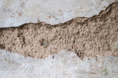 Hasarlı gri beton bir duvarın yakın çekimi. Geniş yatay bir çatlak. İçindeki kaba, kumlu alçı tabakayı ortaya çıkarıyor..
